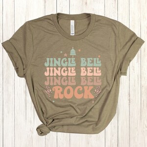 May include: A green t-shirt with the text "Jingle Bell Jingle Bell Jingle Bell Rock" in a retro font. The text is in shades of pink, blue, and green. There are small stars and a lightning bolt with a leopard print design.