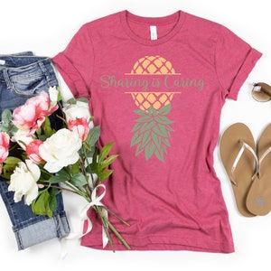 Puede incluir: Una camiseta rosa con un gr&aacute;fico de pi&ntilde;a y el texto "Sharing is Caring". La camiseta est&aacute; colocada sobre un fondo blanco con un par de pantalones vaqueros azules, un ramo de flores rosas y blancas y un par de chanclas marrones.
