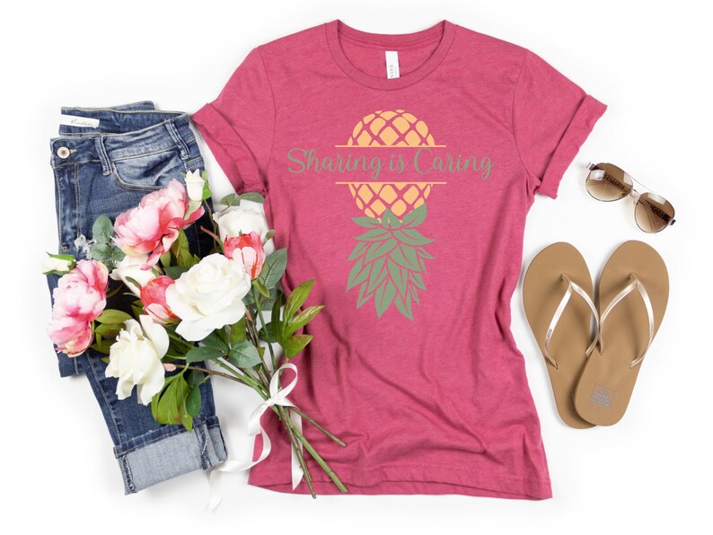 Puede incluir: Una camiseta rosa con un gr&aacute;fico de pi&ntilde;a y el texto "Sharing is Caring". La camiseta est&aacute; colocada sobre un fondo blanco con un par de pantalones vaqueros azules, un ramo de flores rosas y blancas y un par de chanclas marrones.