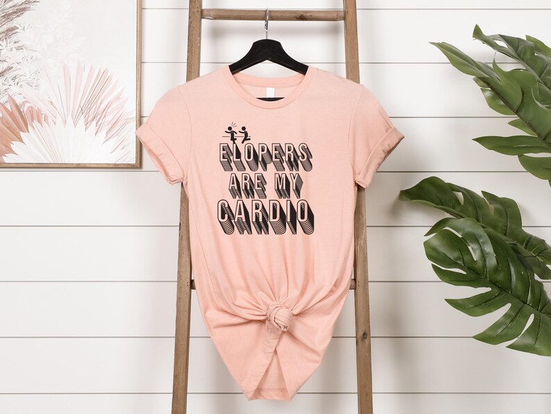 May include: A peach-colored t-shirt with a black graphic that reads "Elopers are my cardio" with a stick figure couple running away.