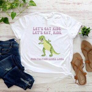 May include: White t-shirt featuring a green dinosaur graphic and the text "Let's eat kids. Let's eat, kids. Punctuation saves lives."