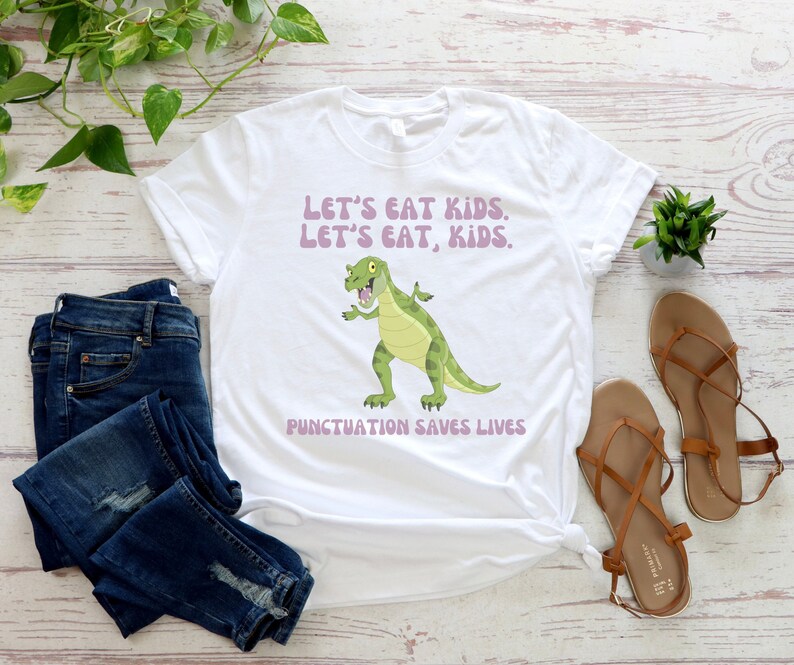 May include: White t-shirt featuring a green dinosaur graphic and the text "Let's eat kids. Let's eat, kids. Punctuation saves lives."