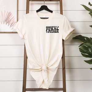 Peut inclure: Un t-shirt cr&egrave;me avec le texte "Somebody's Feral Teacher" en lettres noires.