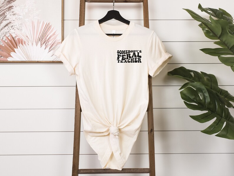 Peut inclure: Un t-shirt cr&egrave;me avec le texte "Somebody's Feral Teacher" en lettres noires.