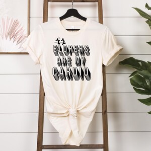 May include: A white t-shirt with black text that reads "ELOPERS ARE MY CARDIO" in a retro font. The text is layered and has a 3D effect. There are two stick figures running in the upper left corner of the text.