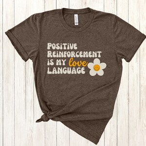 Puede incluir: Una camiseta marr&oacute;n con el texto "Positive Reinforcement is my love language" en una fuente retro con una margarita blanca con una carita sonriente amarilla.