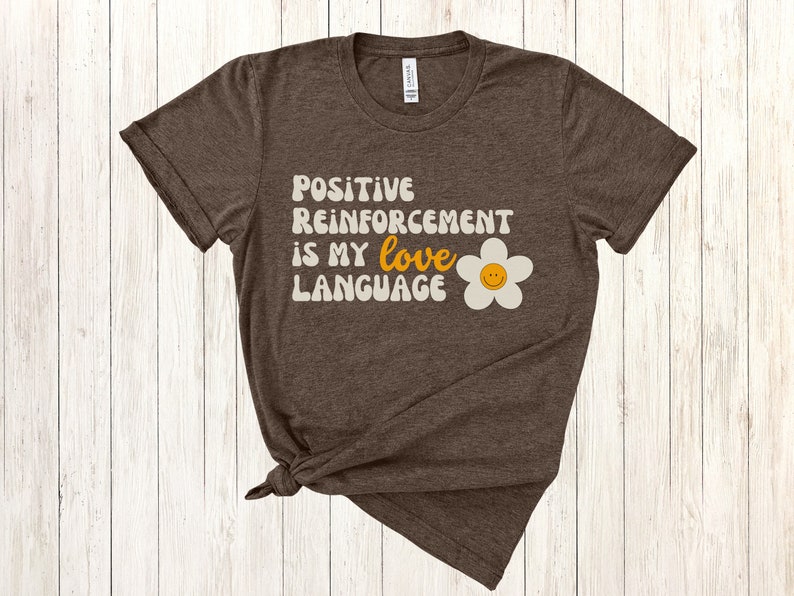 Puede incluir: Una camiseta marr&oacute;n con el texto "Positive Reinforcement is my love language" en una fuente retro con una margarita blanca con una carita sonriente amarilla.