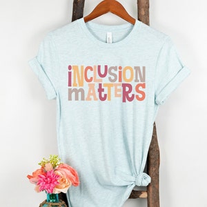 Puede incluir: Una camiseta azul claro con el texto "inclusion matters" impreso en letras de colores. Las letras son en tonos de rosa, naranja y amarillo.