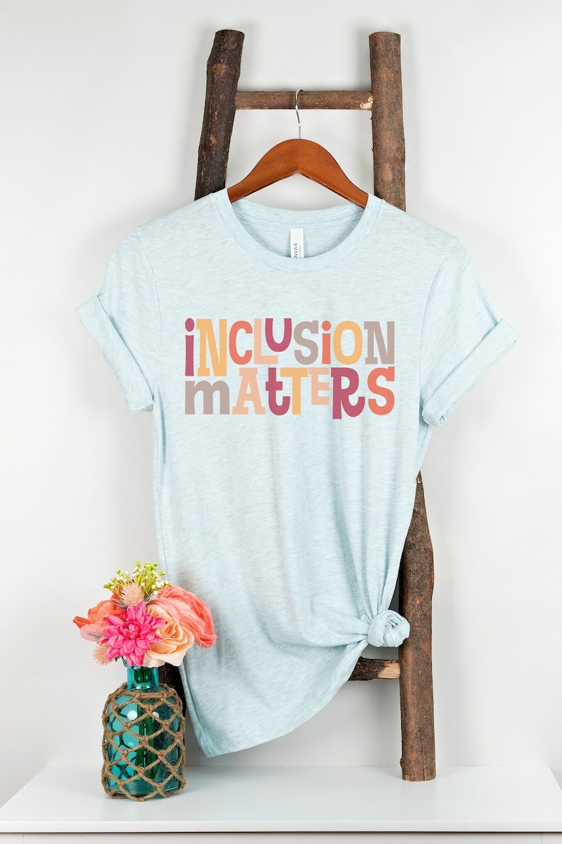 Puede incluir: Una camiseta azul claro con el texto "inclusion matters" impreso en letras de colores. Las letras son en tonos de rosa, naranja y amarillo.