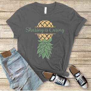 Puede incluir: Una camiseta gris oscuro con un gr&aacute;fico de pi&ntilde;a y el texto "Sharing is Caring" impreso en la parte delantera.
