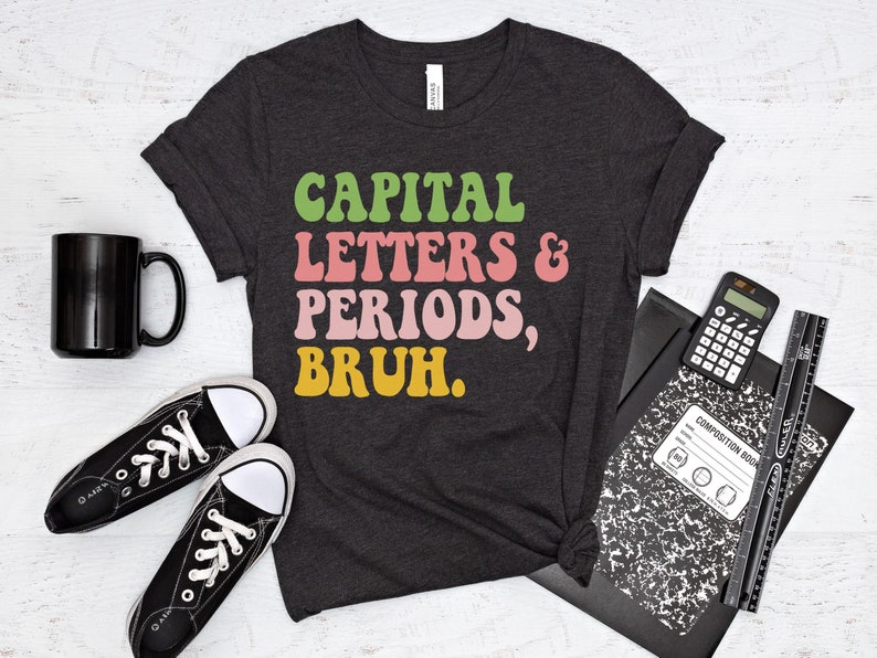 Puede incluir: Una camiseta gris oscuro con el texto "CAPITAL LETTERS & PERIODS, BRUH." en letras retro de colores.