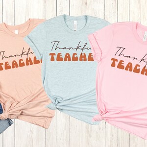 Puede incluir: Tres camisetas con el texto "Thankful Teacher" en una fuente retro. Las camisetas son de color rosa, azul claro y melocot&oacute;n.