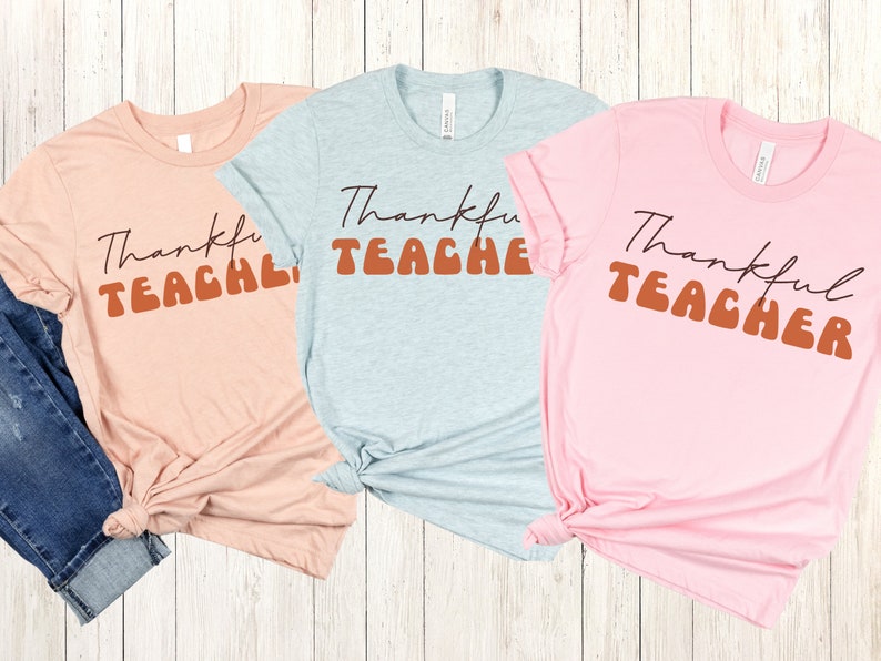 Puede incluir: Tres camisetas con el texto "Thankful Teacher" en una fuente retro. Las camisetas son de color rosa, azul claro y melocot&oacute;n.