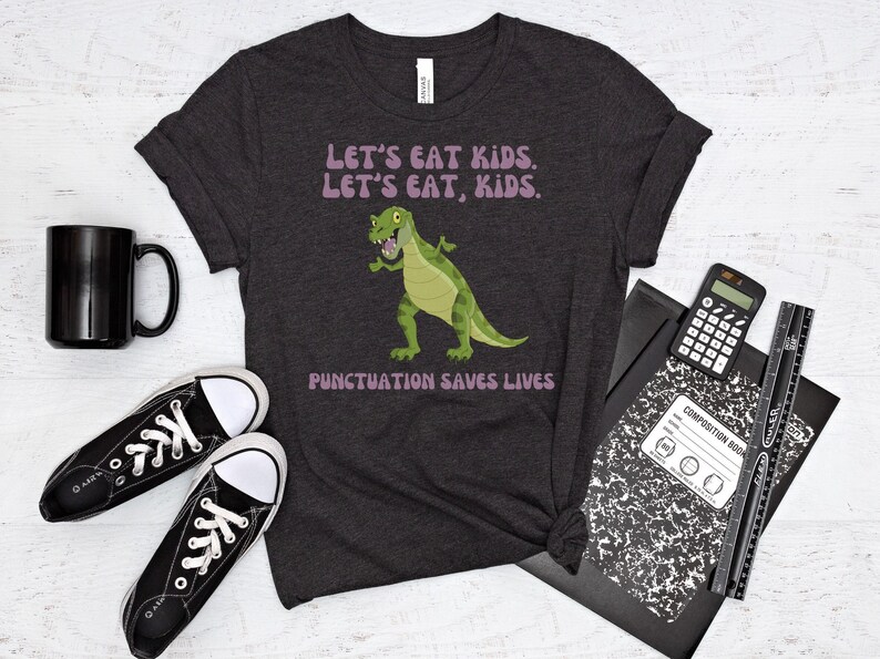 May include: A dark grey t-shirt featuring a green dinosaur graphic and the text "Let's eat kids. Let's eat, kids. Punctuation saves lives." in purple.