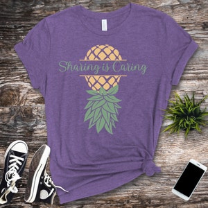 Puede incluir: Camiseta morada con un gr&aacute;fico de pi&ntilde;a y el texto "Sharing is Caring" impreso en la parte delantera.