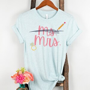 May include: A light blue t-shirt with the text "ms. mrs." in pink lettering. The word "ms." is crossed out with a black line and a pencil is drawn above the line. A diamond ring is drawn below the text.