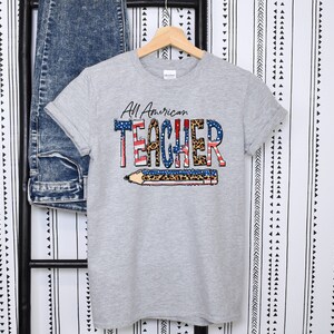 May include: Gray t-shirt with a graphic print of a pencil with an American flag design and the text "All American TEACHER" in black and leopard print.