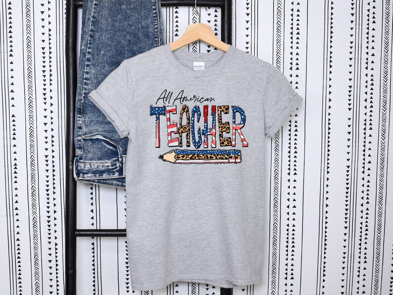 May include: Gray t-shirt with a graphic print of a pencil with an American flag design and the text "All American TEACHER" in black and leopard print.