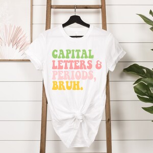 Puede incluir: Camiseta blanca con el texto "CAPITAL LETTERS & PERIODS, BRUH." en una fuente retro colorida.