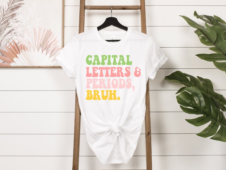 Puede incluir: Camiseta blanca con el texto "CAPITAL LETTERS & PERIODS, BRUH." en una fuente retro colorida.