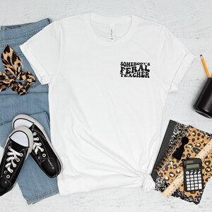 Peut inclure: T-shirt blanc avec le texte "Somebody's Feral Teacher" en noir. Le t-shirt est pos&eacute; sur une surface blanche avec une paire de baskets noires, un jean bleu, un chouchou &agrave; cheveux &agrave; imprim&eacute; l&eacute;opard, un carnet &agrave; imprim&eacute; l&eacute;opard, une tasse noire, des crayons, une r&egrave;gle et une calculatrice.