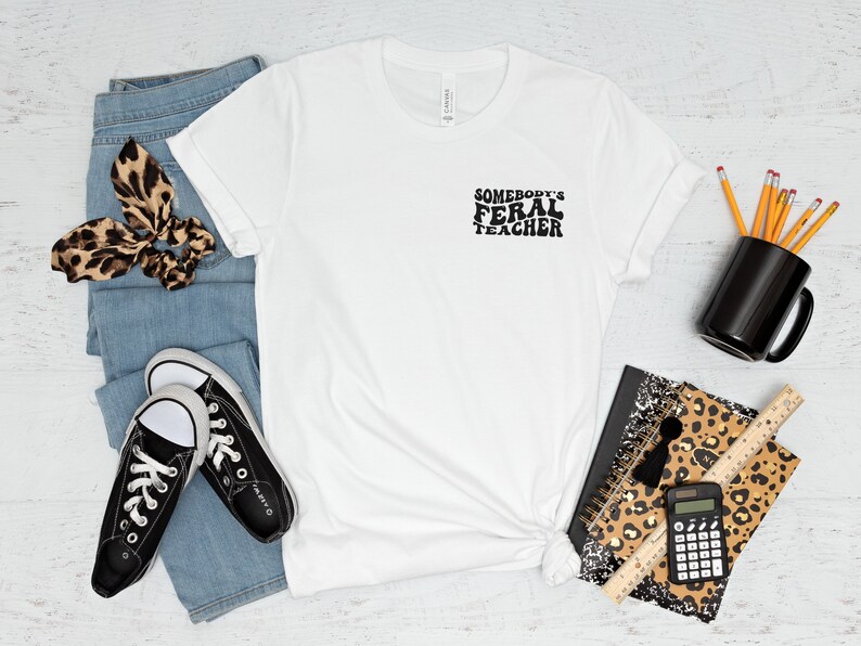Peut inclure: T-shirt blanc avec le texte "Somebody's Feral Teacher" en noir. Le t-shirt est pos&eacute; sur une surface blanche avec une paire de baskets noires, un jean bleu, un chouchou &agrave; cheveux &agrave; imprim&eacute; l&eacute;opard, un carnet &agrave; imprim&eacute; l&eacute;opard, une tasse noire, des crayons, une r&egrave;gle et une calculatrice.