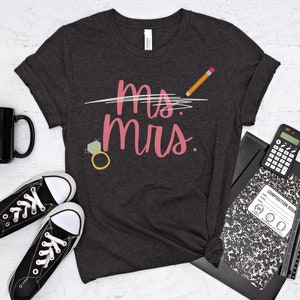 May include: A dark gray t-shirt with the text "Ms. crossed out and Mrs. written in pink. A pencil is drawn above the text. A diamond ring with a sparkly stone is drawn below the text.