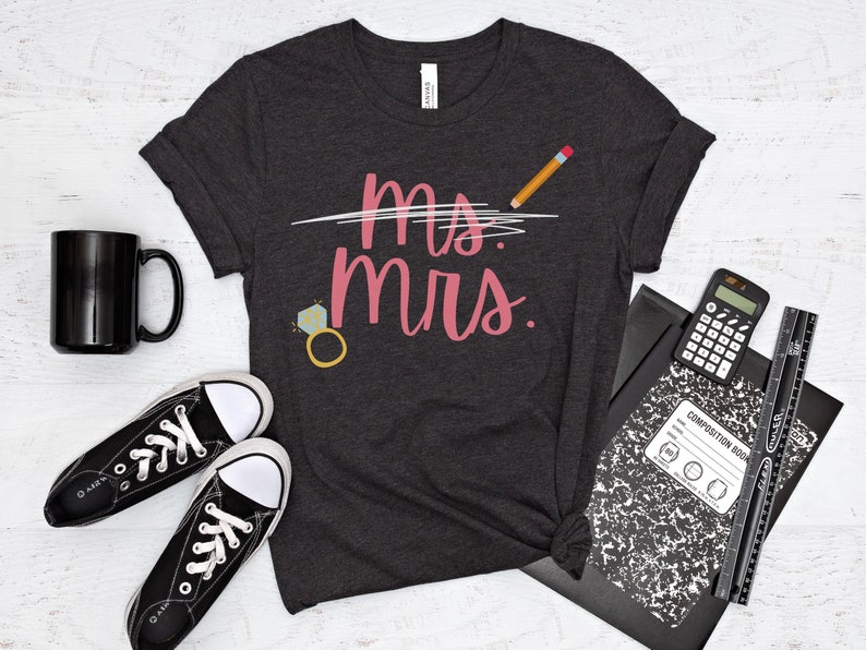May include: A dark gray t-shirt with the text "Ms. crossed out and Mrs. written in pink. A pencil is drawn above the text. A diamond ring with a sparkly stone is drawn below the text.