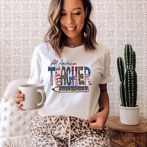 May include: White t-shirt with a red, white, and blue American flag design and the text "All American Teacher" with a pencil graphic. The shirt is being worn by a person with brown hair and a leopard print pajama pants.