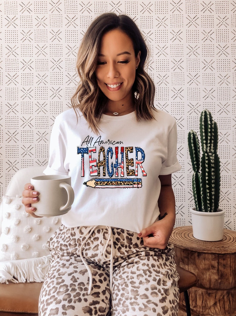 May include: White t-shirt with a red, white, and blue American flag design and the text "All American Teacher" with a pencil graphic. The shirt is being worn by a person with brown hair and a leopard print pajama pants.