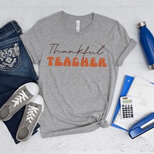 Puede incluir: Una camiseta gris con el texto "Thankful Teacher" en letras naranja y marr&oacute;n. La camiseta est&aacute; colocada sobre una superficie de madera blanca con un par de pantalones vaqueros azules, una botella de agua, una calculadora, una grapadora y un cuaderno.
