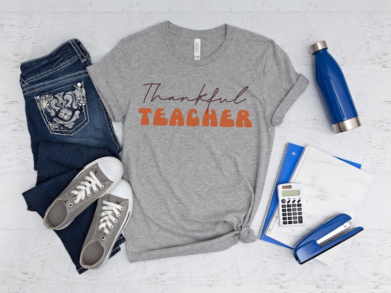 Puede incluir: Una camiseta gris con el texto "Thankful Teacher" en letras naranja y marr&oacute;n. La camiseta est&aacute; colocada sobre una superficie de madera blanca con un par de pantalones vaqueros azules, una botella de agua, una calculadora, una grapadora y un cuaderno.