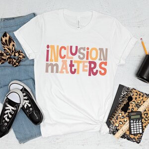 Puede incluir: Camiseta blanca con el texto "inclusion matters" en letras de bloque de colores. La camiseta est&aacute; colocada sobre una superficie de madera blanca con jeans, zapatillas, un porta l&aacute;pices, una libreta, una calculadora y una regla.