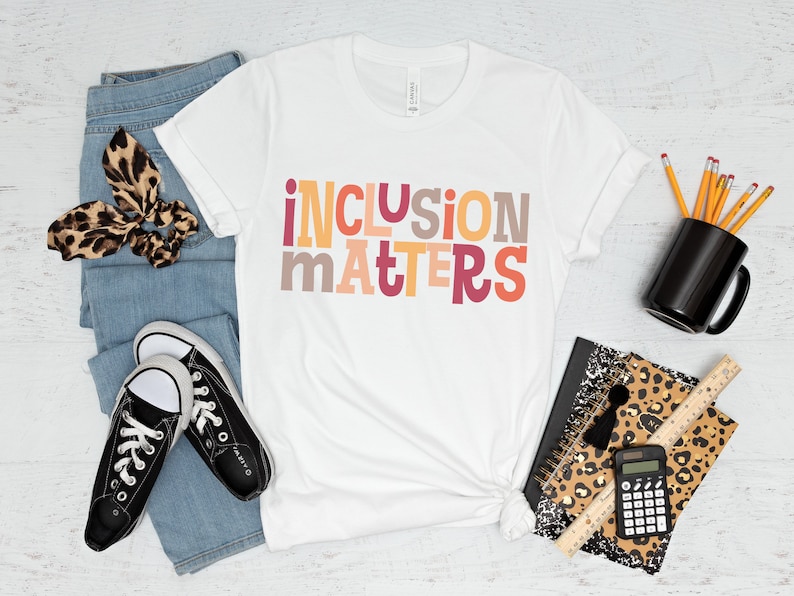 Puede incluir: Camiseta blanca con el texto "inclusion matters" en letras de bloque de colores. La camiseta est&aacute; colocada sobre una superficie de madera blanca con jeans, zapatillas, un porta l&aacute;pices, una libreta, una calculadora y una regla.