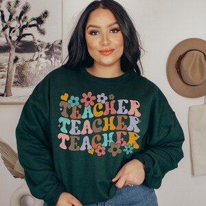 May include: A dark green sweatshirt with a retro-style graphic that says "Teacher Teacher Teacher" in colorful letters and flowers.