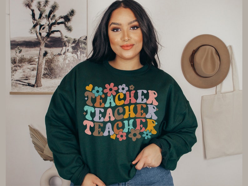 May include: A dark green sweatshirt with a retro-style graphic that says "Teacher Teacher Teacher" in colorful letters and flowers.