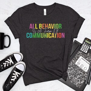 Op de afbeelding: Een donkergrijs T-shirt met de tekst "ALL BEHAVIOR IS A FORM OF COMMUNICATION" in regenboogkleuren.