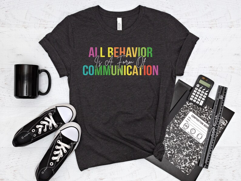 Op de afbeelding: Een donkergrijs T-shirt met de tekst "ALL BEHAVIOR IS A FORM OF COMMUNICATION" in regenboogkleuren.