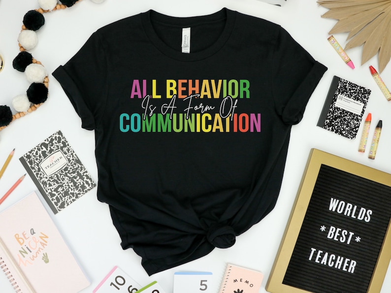 Op de afbeelding: Zwart T-shirt met de tekst "ALL BEHAVIOR Is A Form Of COMMUNICATION" in regenboogkleuren.
