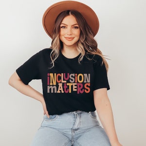 Puede incluir: Una mujer lleva una camiseta negra con el texto "inclusion matters" en letras de colores. La camiseta tiene mangas cortas y un corte holgado. Lleva un sombrero marr&oacute;n y unos vaqueros azules.