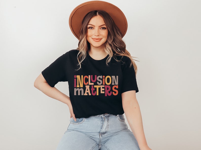 Puede incluir: Una mujer lleva una camiseta negra con el texto "inclusion matters" en letras de colores. La camiseta tiene mangas cortas y un corte holgado. Lleva un sombrero marr&oacute;n y unos vaqueros azules.