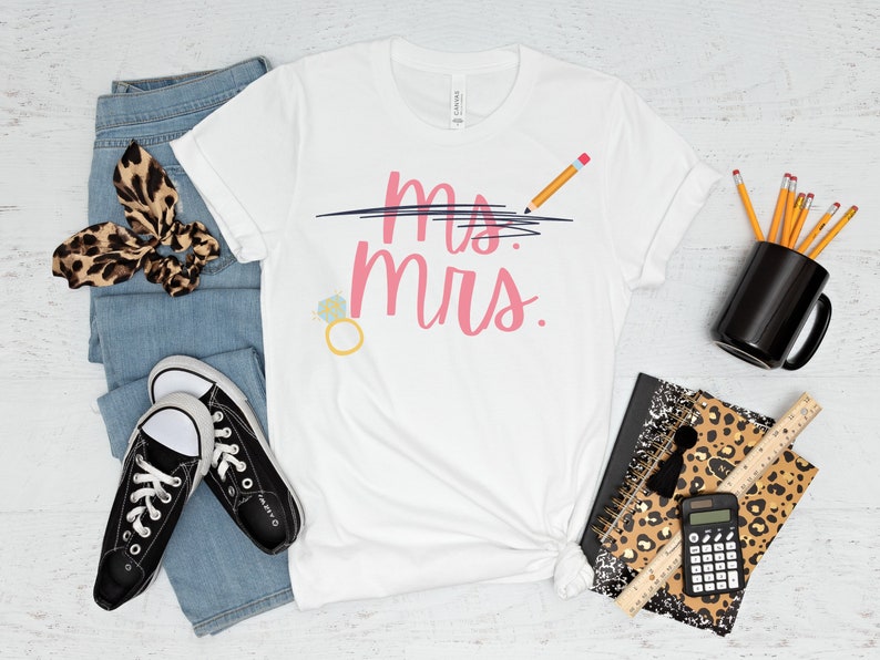 May include: White t-shirt with the text "Mrs." crossed out and "Mrs." written in pink below. A pencil is drawn above the crossed out text. A ring with a diamond is drawn to the left of the text.