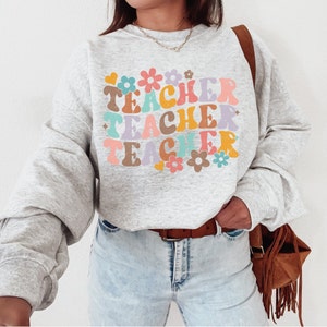 May include: A gray sweatshirt with the word "TEACHER" repeated three times in a retro font with colorful flowers and hearts.