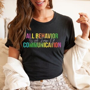 Op de afbeelding: Een zwart T-shirt met de tekst "ALL BEHAVIOR Is A Form Of COMMUNICATION" in regenboogkleuren.