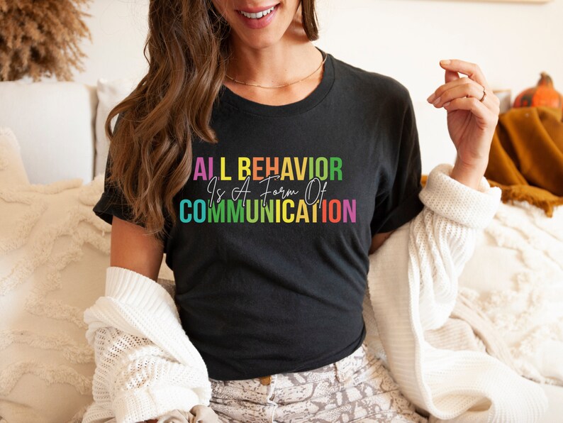 Op de afbeelding: Een zwart T-shirt met de tekst "ALL BEHAVIOR Is A Form Of COMMUNICATION" in regenboogkleuren.