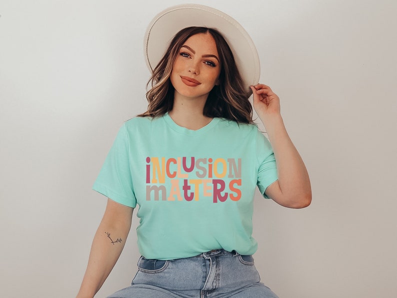 Puede incluir: Una mujer lleva una camiseta azul claro con el texto "inclusion matters" en letras de colores. Las letras son de diferentes colores, incluyendo rosa, amarillo, naranja y gris. Lleva un sombrero blanco y unos vaqueros azules.