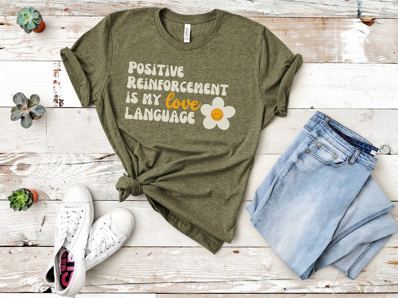 Puede incluir: Una camiseta verde con el texto "Positive Reinforcement is my love language" y una margarita amarilla con una cara sonriente.