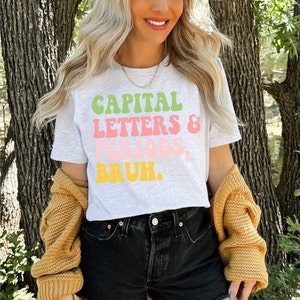 Puede incluir: Una camiseta blanca con un gr&aacute;fico que dice "CAPITAL LETTERS & PERIODS, BRUH." en letras retro de colores.