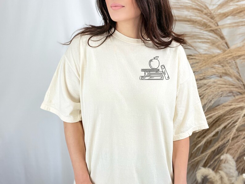 May include: A white t-shirt with a black and white graphic of an apple on top of a stack of books.
