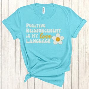 Puede incluir: Una camiseta azul turquesa con el texto "Positive Reinforcement is my love language" en una fuente retro con una margarita blanca con una carita sonriente amarilla.